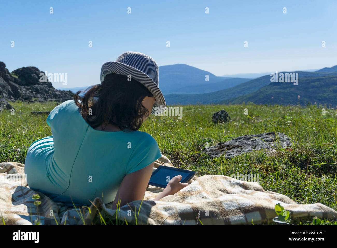 Ragazza in abito e hat seduta su una coperta con una compressa nelle sue mani su un prato alpino contro uno sfondo di montagne, la vista dal retro Foto Stock