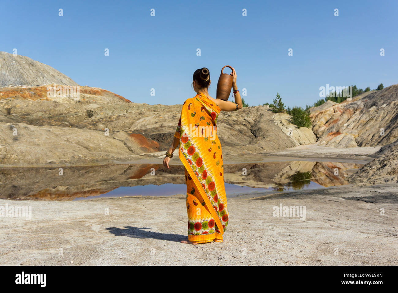 Giovane donna in un folk vestito con un vaso di creta sulla sua spalla va a una molla in una zona arida Foto Stock