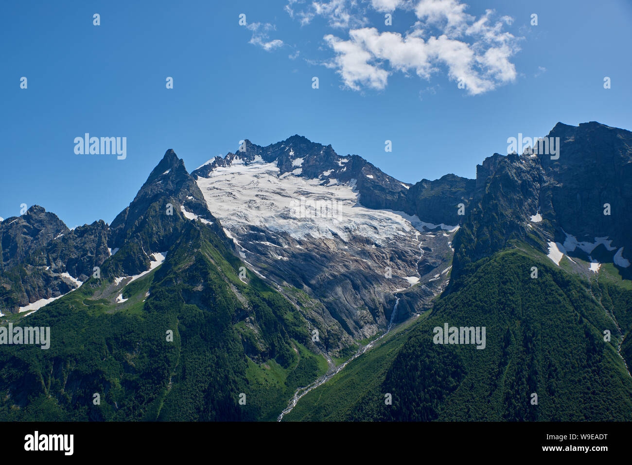 Picchi di montagna con un ghiacciaio e l inizio di un ruscello di montagna che scorre attraverso una foresta di pini. Caucaso del Nord, Russia Foto Stock