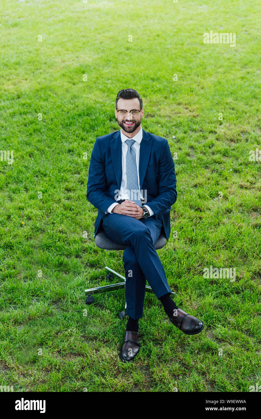 Angolo di alta vista di felice giovane imprenditore seduto nella sedia per ufficio nel parco e loooking in telecamera Foto Stock