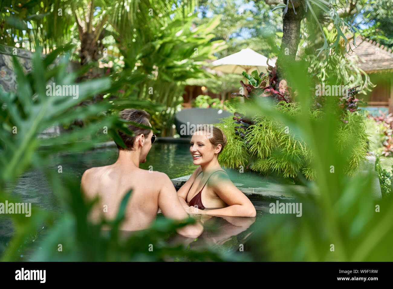 Unposed colpo di coppia felice in piedi insieme nella lussureggiante resort pool di hotel di lusso in tropicale Bali durante le vacanze Foto Stock