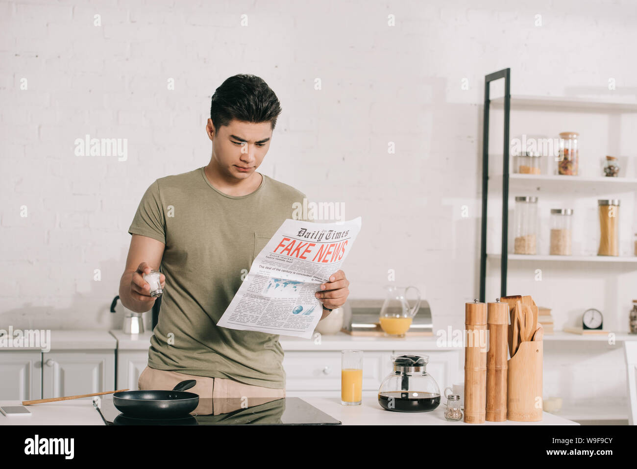 Bel uomo asiatico prepara la colazione durante la lettura fake news giornale in cucina Foto Stock