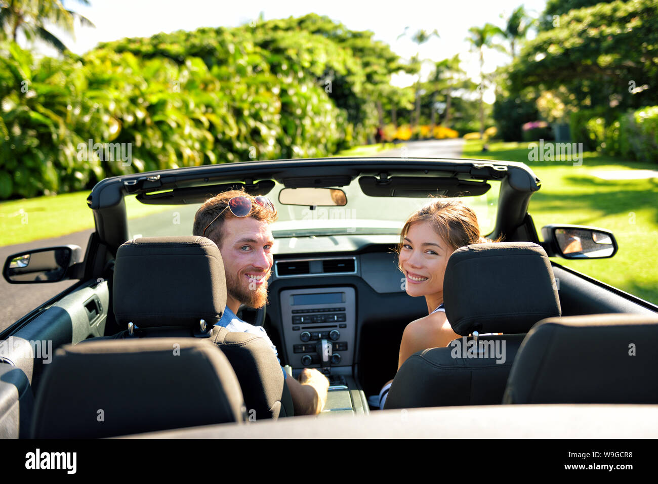 Coppia felice in auto sulla strada estiva di viaggio. Multirazziale coppia giovane spensierate vacanze sulla guida di una cabriolet convertibile automobile sulla carreggiata in città guardando indietro alla fotocamera. Foto Stock