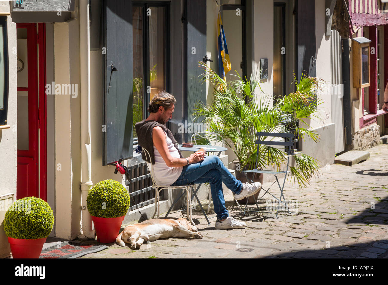 Un uomo si siede e fuma una sigaretta a un tavolo nella sushine su una strada di ciottoli in Honfleur, Normandia, Francia come il suo cane dorme ai suoi piedi Foto Stock