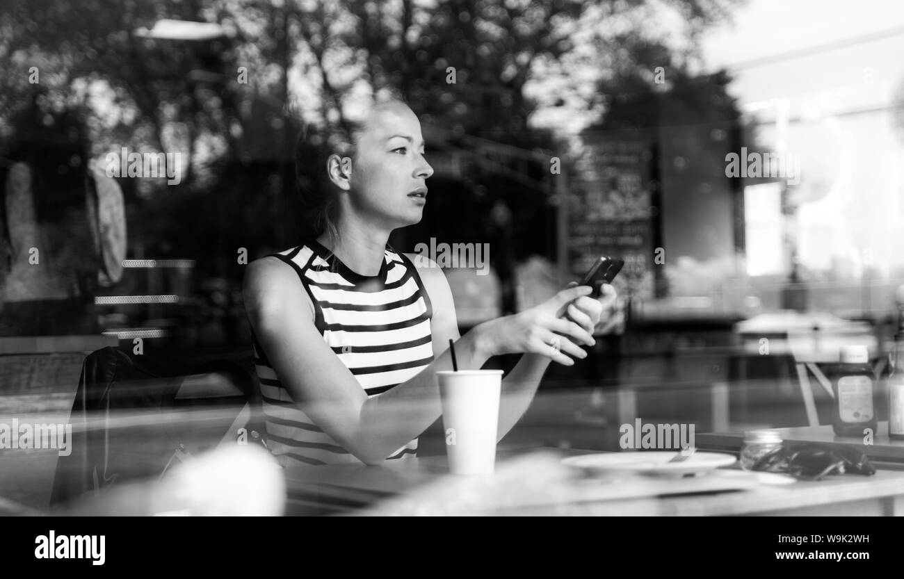 Premurosa donna caucasica tenendo il telefono cellulare mentre si cerca attraverso la caffetteria finestra durante la pausa caffè. Foto Stock
