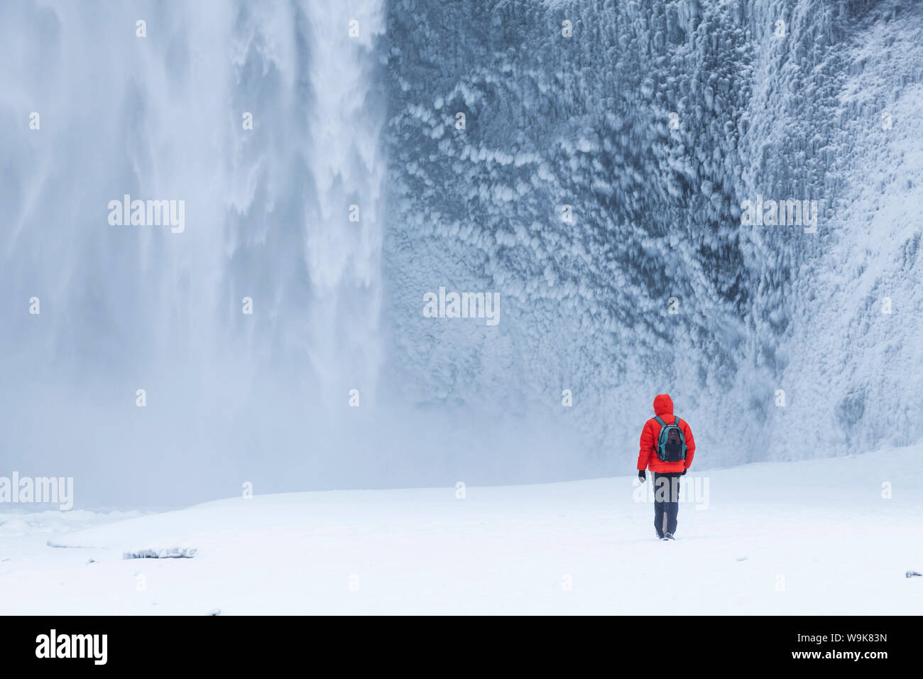 Una persona in giacca rossa a piedi nella neve verso la cascata Skogafoss in inverno, Skogar, Sud Islanda, Islanda, regioni polari Foto Stock