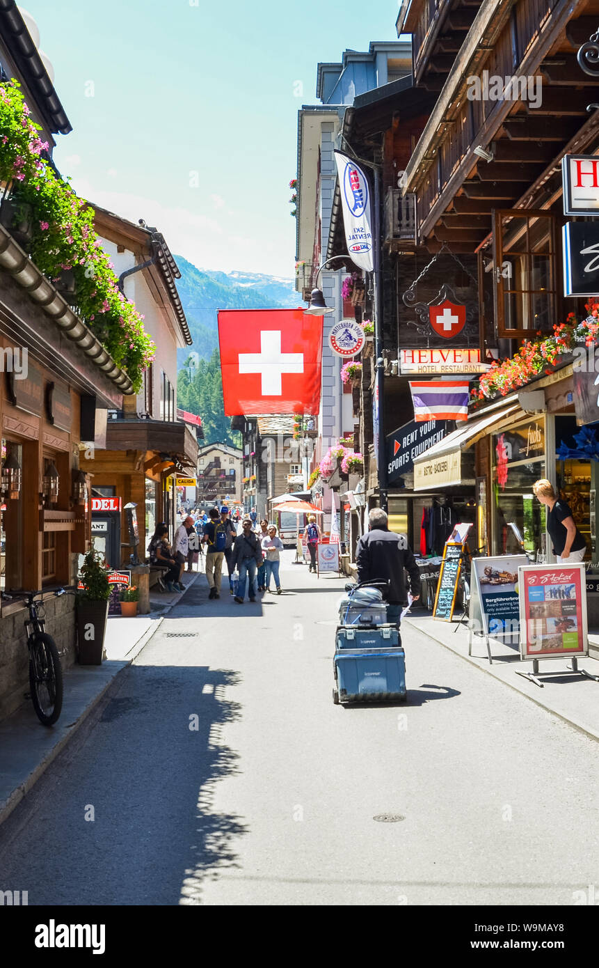 Zermatt, Svizzera - 10 Luglio 2019: strada in località alpina Zermatt durante la stagione estiva. Sventola bandiera svizzera. Negozi e ristoranti su una strada. Turi Foto Stock