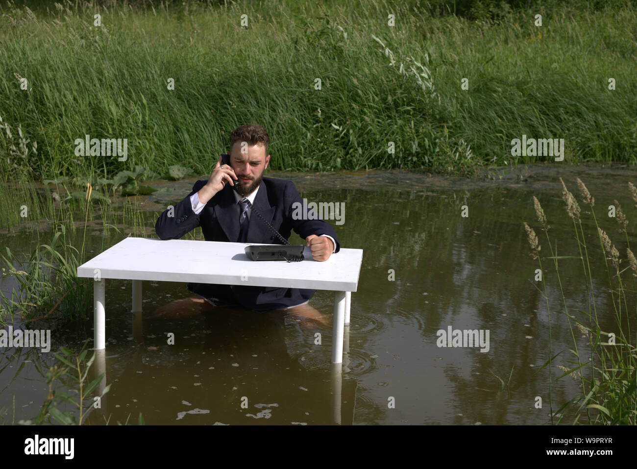 Un uomo barbuto con un entusiasta, preoccupati di fronte sta parlando al telefono. Egli bussa il pugno sul tavolo. Sedersi nella palude, in acqua. Foto Stock