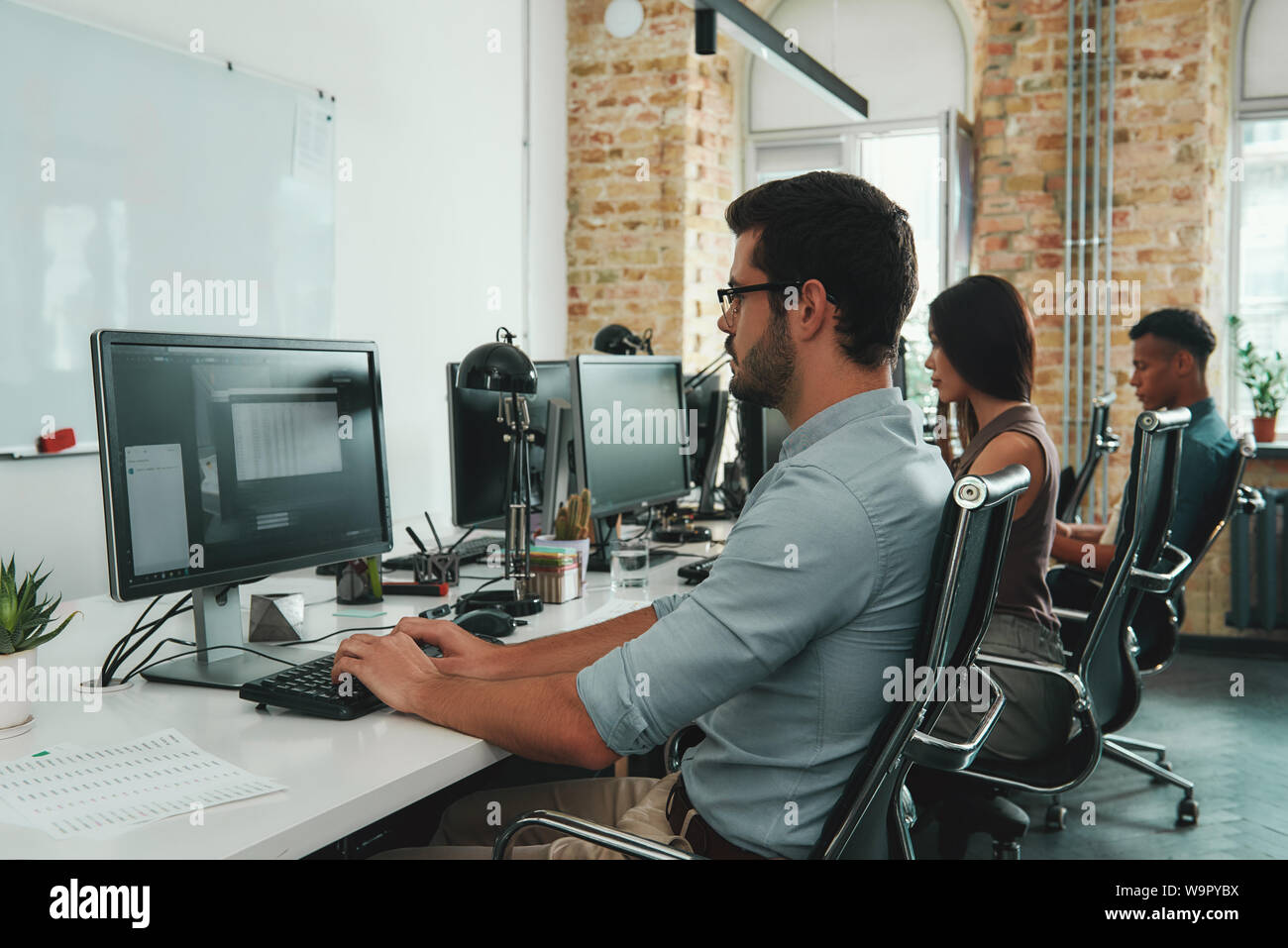 Focalizzati sul lavoro. Vista laterale di giovani occupati i dipendenti che lavorano su computer mentre è seduto alla scrivania in moderno spazio aperto. Il concetto di lavoro. Sul posto di lavoro. Lavoro di squadra Foto Stock