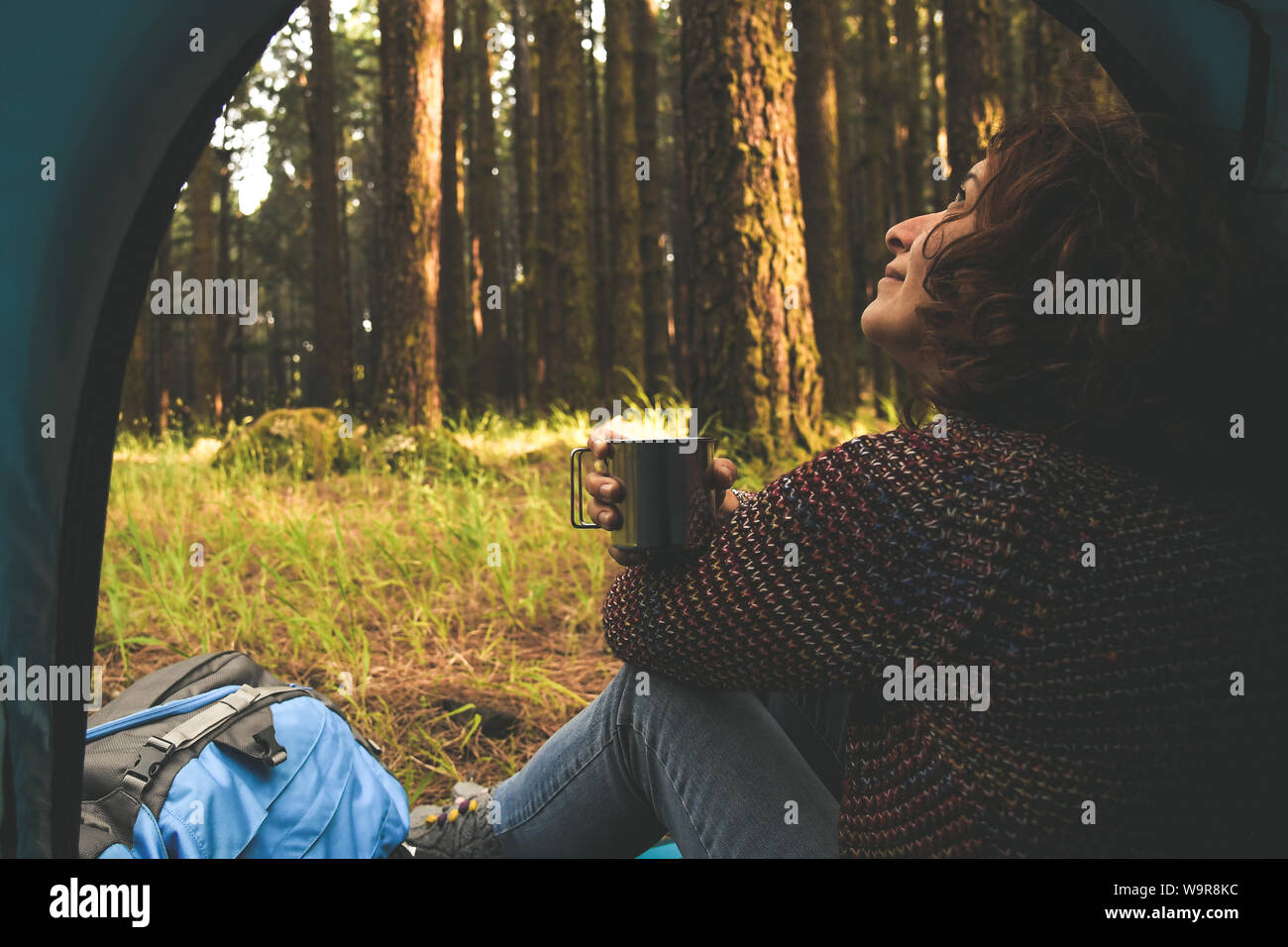 Bella donna seduta nella tenda in cerca della natura selvaggia fuori beve una tazza di caffè alla moda ragazza sola rilassante nella foresta dopo un trekking h Foto Stock