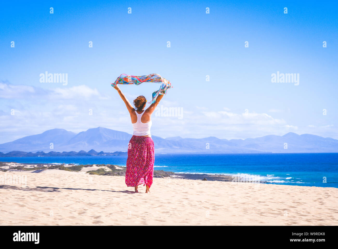 Bella ragazza giovane con mosche sciarpa nelle sue mani oltre le dune di sabbia bianca a Fuerteventura. Colorate vacanza esotici cartolina. Blu oceano e cielo in ba Foto Stock