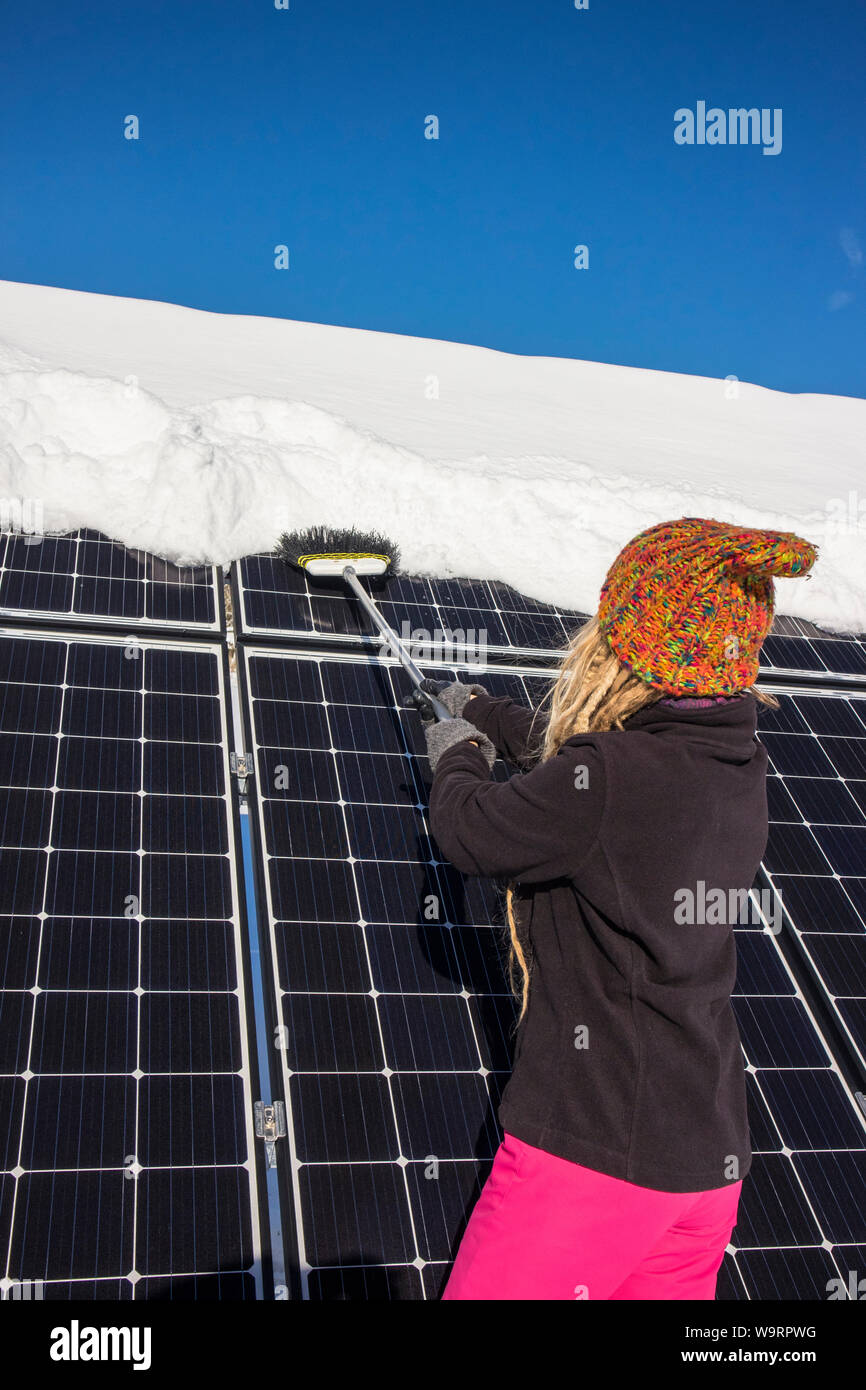 Donna spingendo la neve off pannelli solari in inverno. Se la neve copre i pannelli non sono in grado di produrre energia. Piccolo privato home casa sullo sfondo. Foto Stock