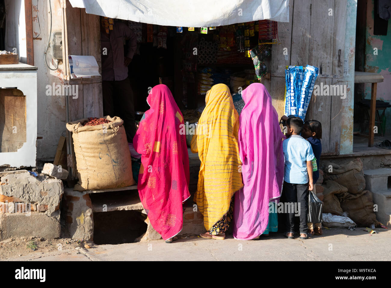 Drei Frauen in bunten sari vor einem Geschäft stehend (Rücken zur Kamera), zwei Kinder stehen neben ihnen. In Straßenszene Mandawa (Rajasthan) in Ind Foto Stock