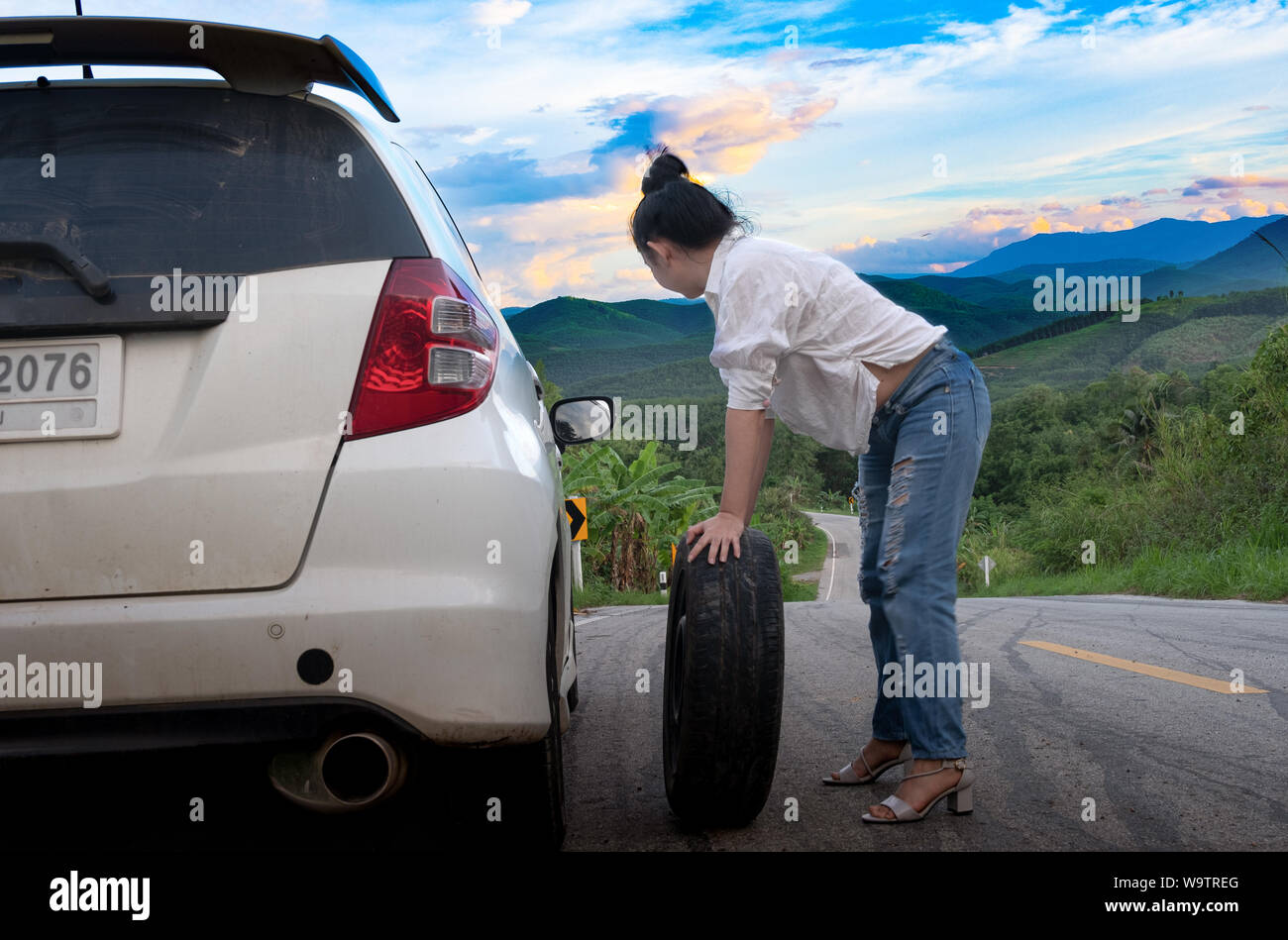 Giovane donna attraente in abbigliamento formale tenendo un ricambio pneumatico auto sulla strada pubblica nella zona forestale in montagna e sullo sfondo del cielo Foto Stock