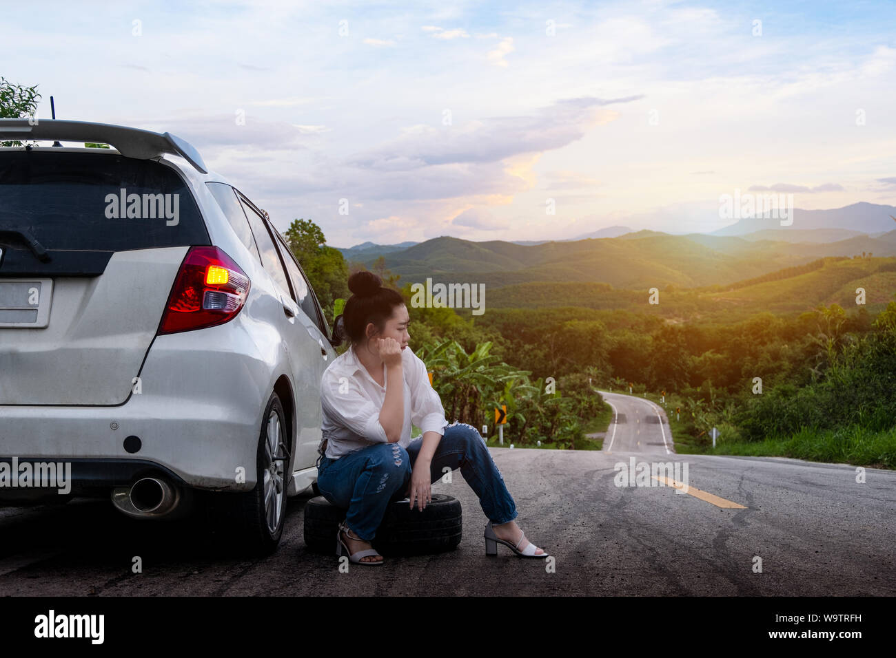 Giovane bella signora sedersi vicino a vettura per chiedere aiuto sulla strada pubblica nella zona forestale in montagna e sullo sfondo del cielo Foto Stock
