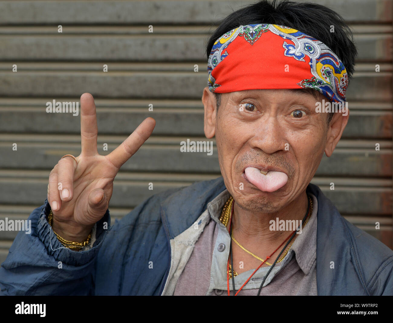 Divertente di mezza età uomo tailandese con una colorata bandana bastoni fuori la sua lingua e visualizza il segno V con la sua mano destra. Foto Stock