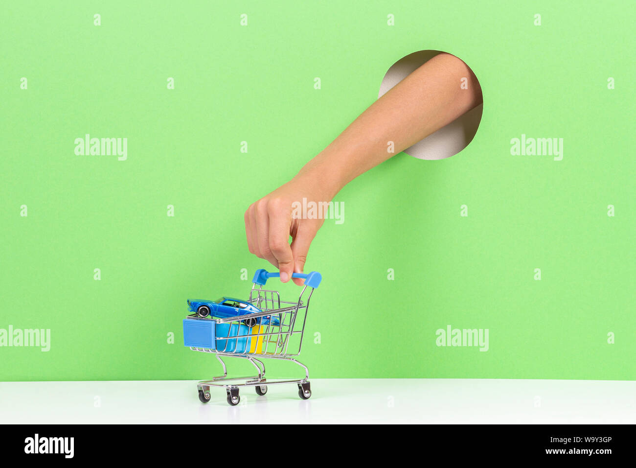 Kid mano toccando un mini carrello della spesa con giocattoli attraverso il foro su sfondo verde chiaro Foto Stock