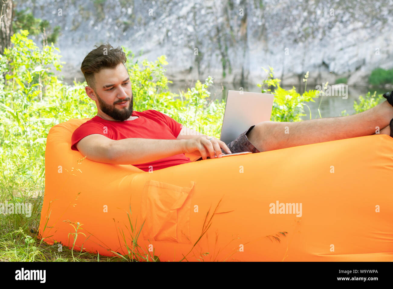 Giovane imprenditore barbuto con laptop sulle ginocchia è seduta sul orange divano gonfiabile sull'erba nella foresta. Foto Stock