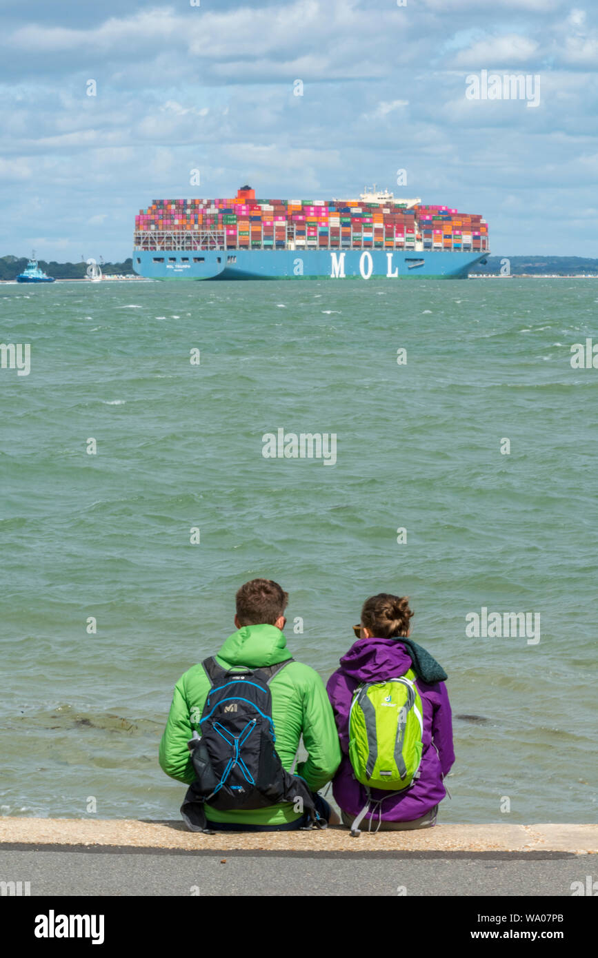 una giovane coppia che indossa giacche impermeabili e zaini seduti su una spiaggia di ciottoli guardando una grande nave container nel solent a cowes. Foto Stock