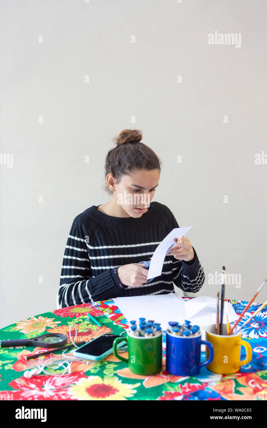 Il giovane talento donna artista seduto a tavola con la tovaglia fiorito fare arte con matite, carta, forbici, lente di ingrandimento, spazzola e vernici Foto Stock