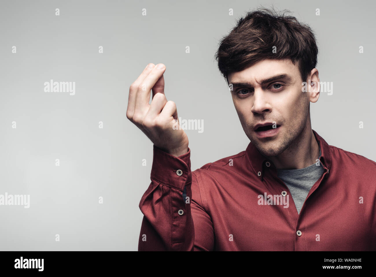 Uomo insoddisfatto che mostra il gesto di denaro e guardando la telecamera isolata sul grigio, emozioni umane e concetto di espressione Foto Stock