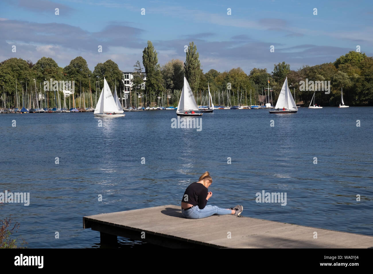 Giovane donna seduta sul molo e guardando la barca a vela gara sull'esterno Lago Alster Amburgo, Germania Foto Stock