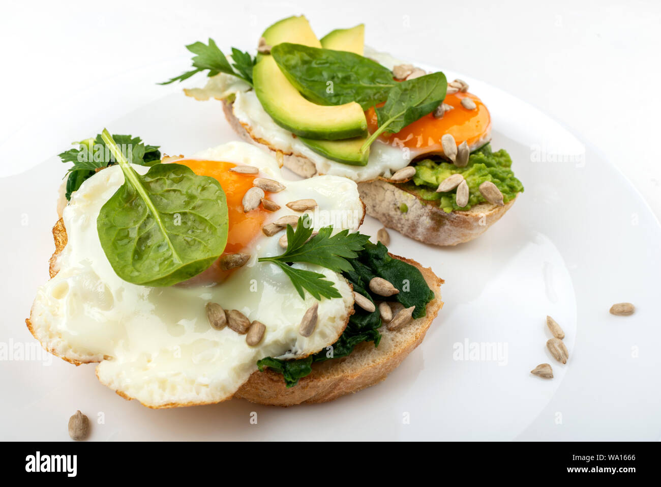 Uova fritte su un tosta pane con avocado, spinaci e semi su una piastra bianca su sfondo bianco Foto Stock