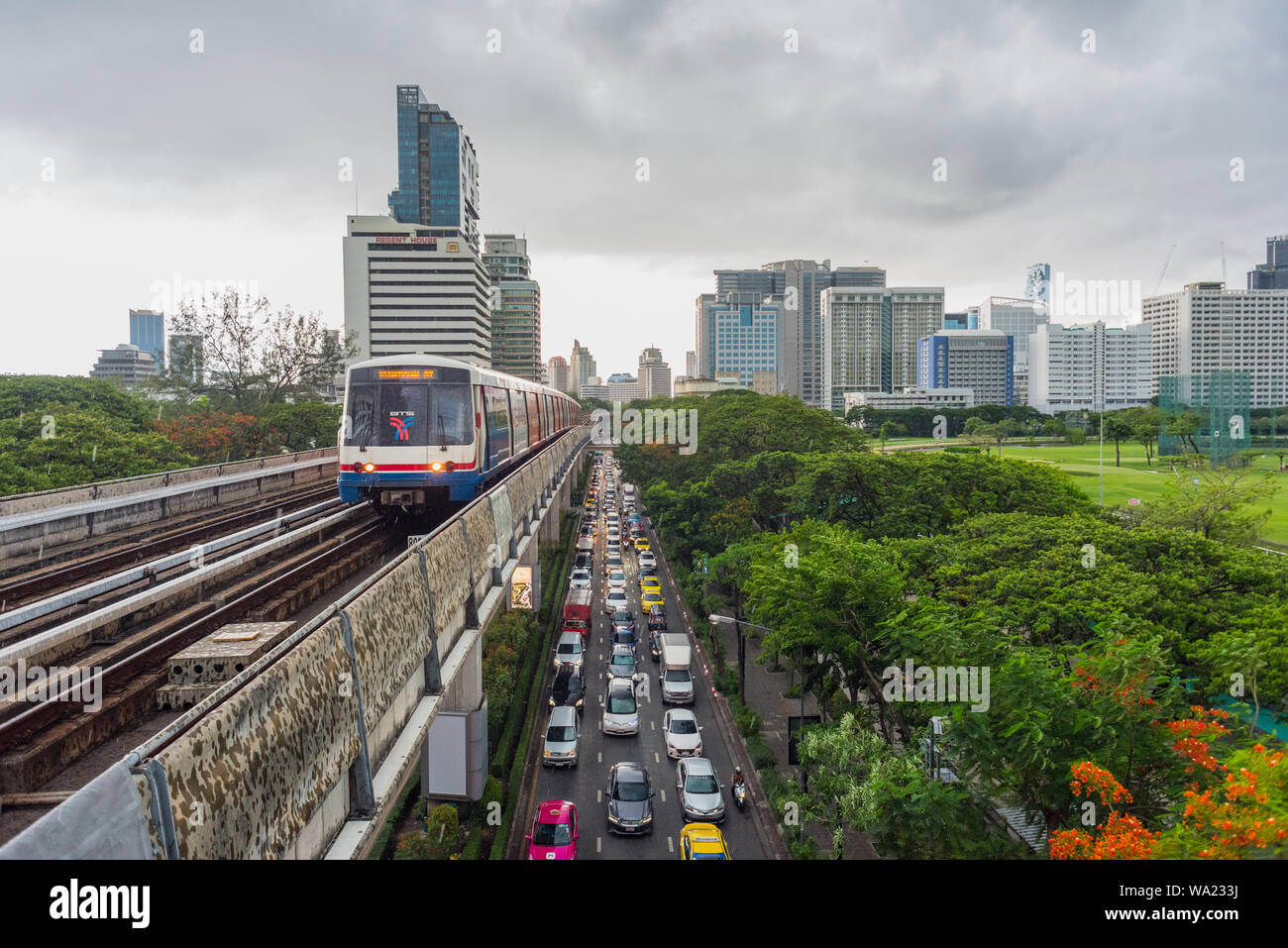 Bangkok - Giugno 9, 2019: un treno arriva a Ratchadamri stazione BTS durante la pioggia, al di sopra del traffico nella strada con il centro cittadino di edifici. Foto Stock