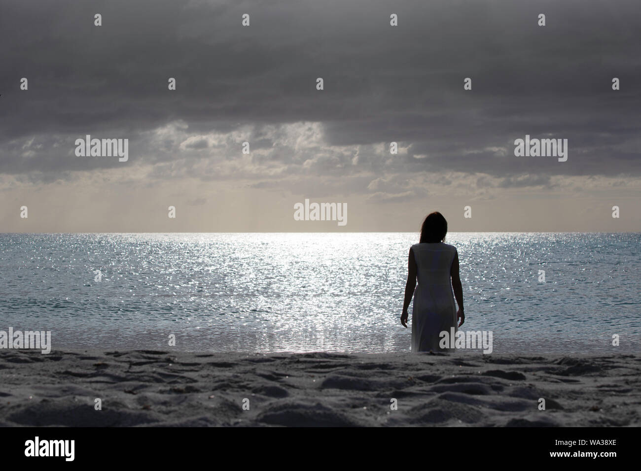 Silhouette di donna da dietro il vestito di una luce abito bianco passeggiate tristemente e premurosamente verso il mare scuro all'alba con drammatica sky Foto Stock