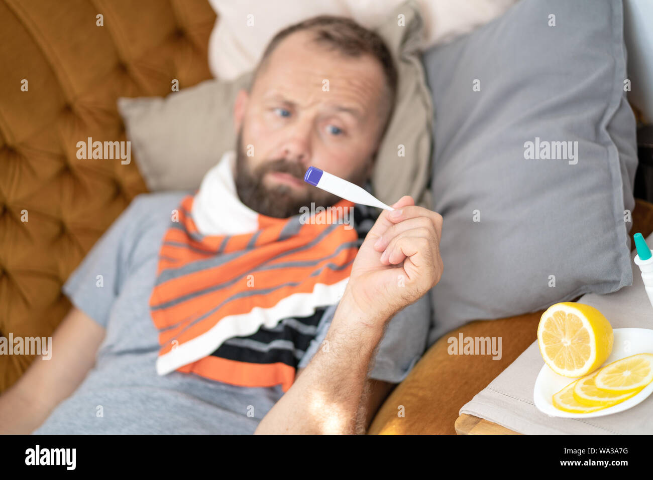 Uomo malato sdraiato sul divano nel controllo della sua temperatura in casa nel salotto. Uomo sciarpa in attesa termometro vicino. Misurare la temperatura. Elevata temperat Foto Stock