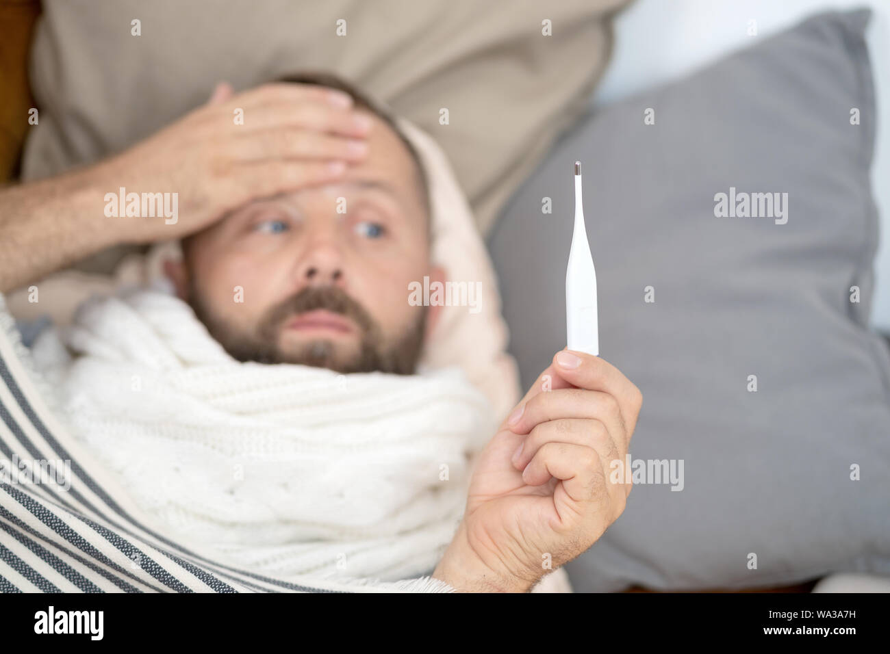 Uomo malato sdraiato sul divano nel controllo della sua temperatura in casa nel salotto. Uomo sciarpa in attesa termometro vicino. Misurare la temperatura. Elevata temperat Foto Stock