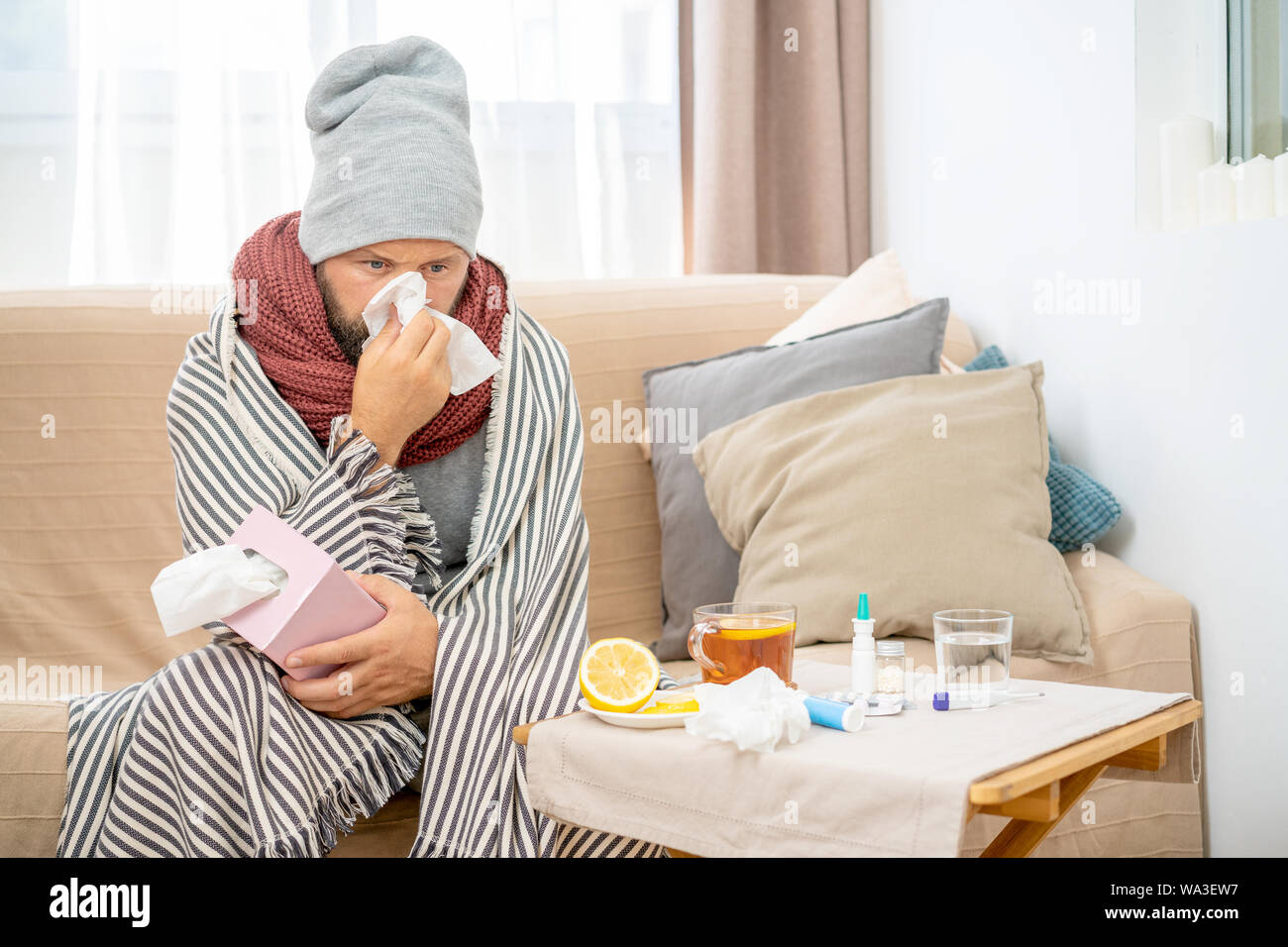 Uomo malato che indossa cappello e sciarpa grigi, che soffia il naso e starnutisce nel tessuto. I maschi hanno influenza, virus o allergia respiratoria. Sano, concetto di medicina Foto Stock