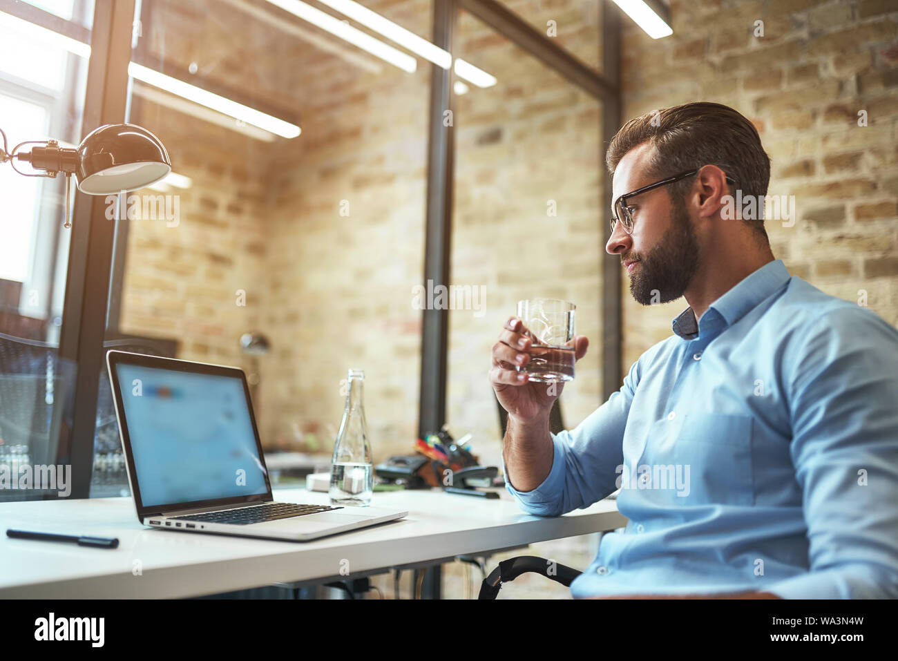 Lavorando sodo. Sete barbuto imprenditore in occhiali e usura formale di bere acqua fresca e guardando il computer portatile mentre seduto in un ufficio moderno. Il concetto di business. Concetto di lavoro Foto Stock