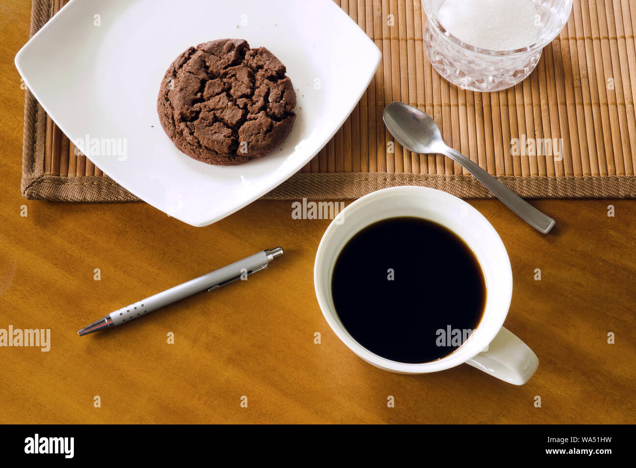 Nero caffè in ceramica tazza bianco, cioccolato cookie su piastra bianca, cucchiaino, penna Bamboo placemat e zucchero-ciotola sul tavolo di legno, indoor, vista dall'alto Foto Stock