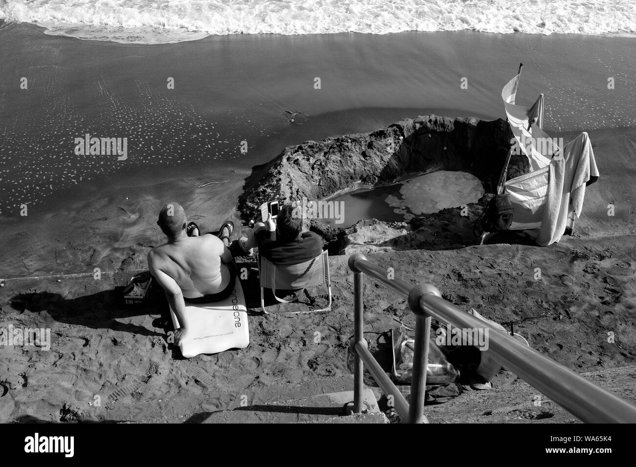 Un giovane seduto sulla spiaggia rilassarsi leggendo un libro e guardando il mare dal litorale sabbioso Foto Stock
