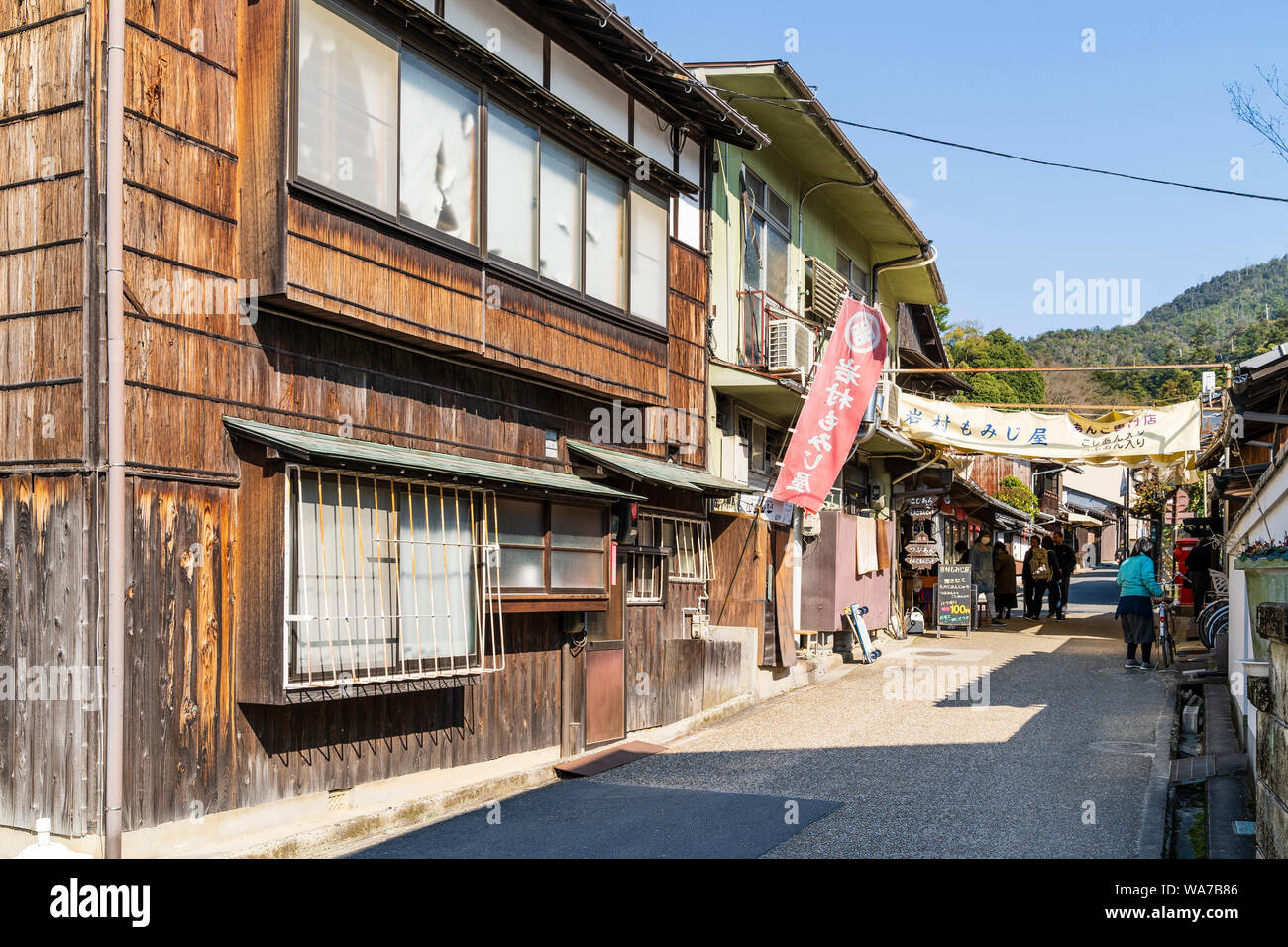 Il Giappone, l'isola di Miyajima. Piccola strada laterale con negozi e case che conduce su per la collina con alcune persone a piedi. Non occupato. Sullo sfondo le montagne della foresta. Foto Stock