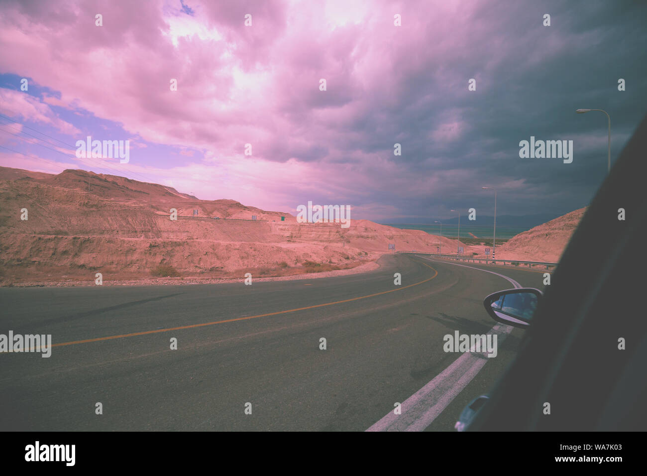 Alla guida di una vettura su una strada di montagna in Israele. Il paesaggio del deserto. Strada vuota. Vista dalla vettura del paesaggio di montagna con drammatica tempestoso Cielo di tramonto. Isr Foto Stock