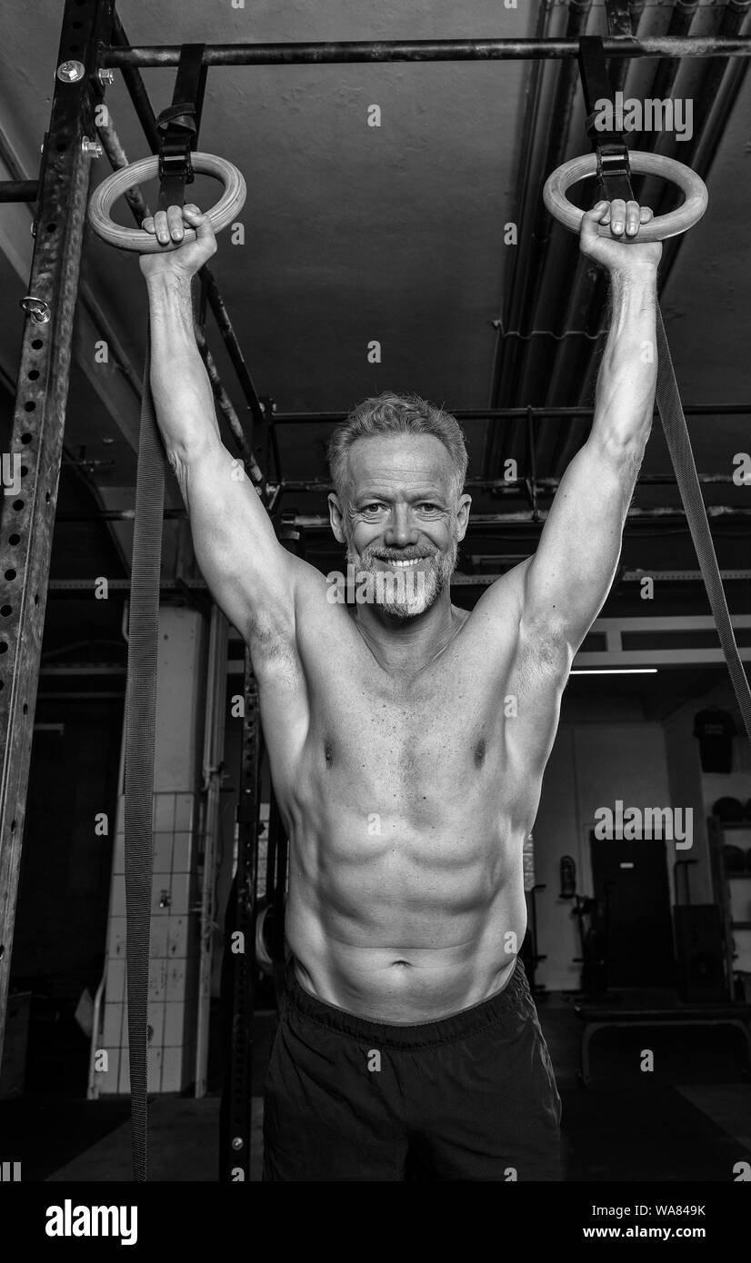 Ritratto di un affascinante vecchio atleta maschio con forti muscoli. L'atletico e sorridente uomo barbuto sta facendo un esercizio con gli anelli. Foto Stock