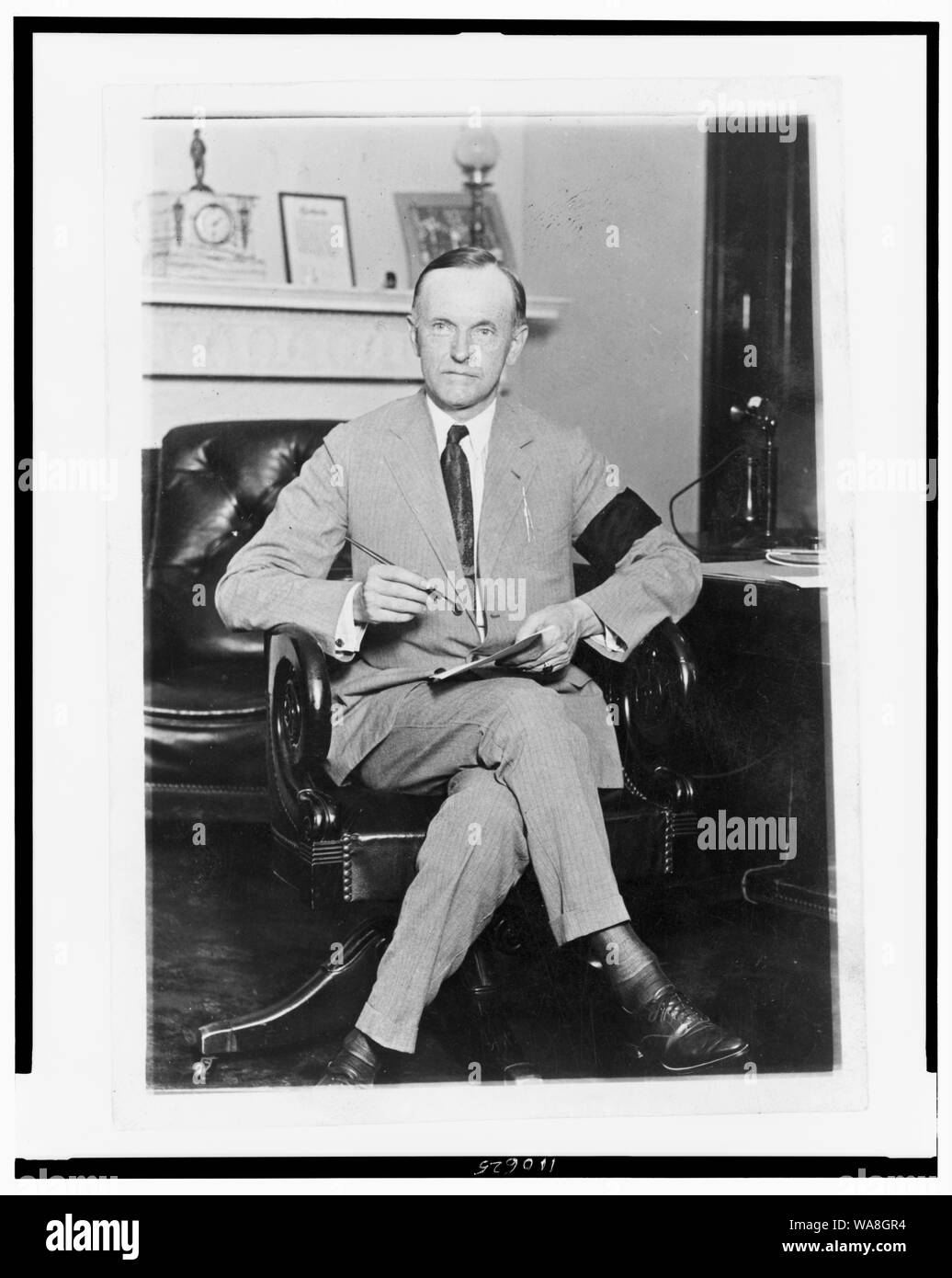 Calvin Coolidge, ritratto a figura intera, seduto alla scrivania, rivolto verso la parte anteriore, tenendo in mano carta e penna, indossa una fascia nera al braccio in lutto per il Presidente Harding Foto Stock