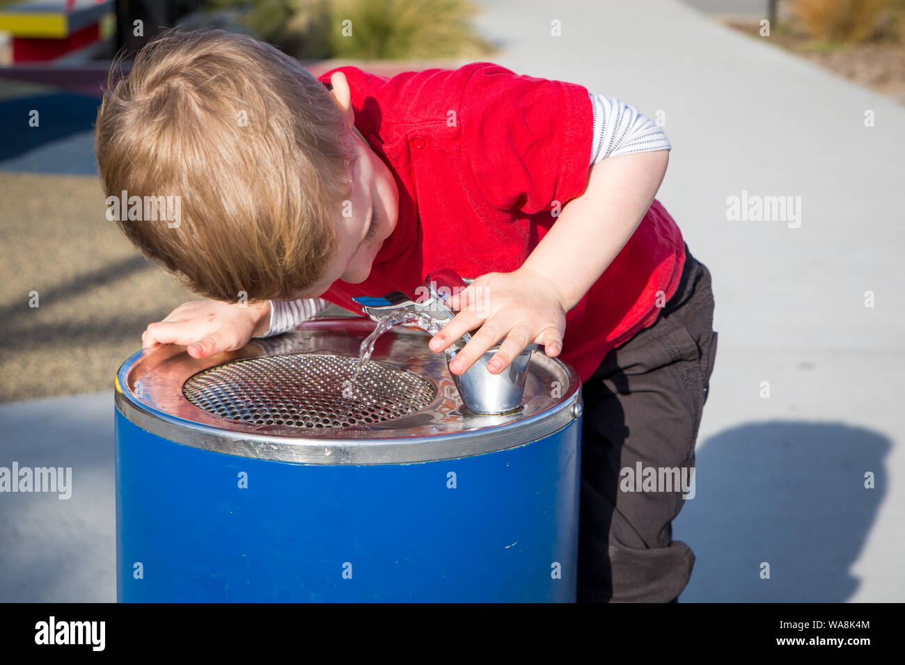 Un piccolo ragazzo beve da una fontana di acqua durante la riproduzione in un parco giochi Foto Stock