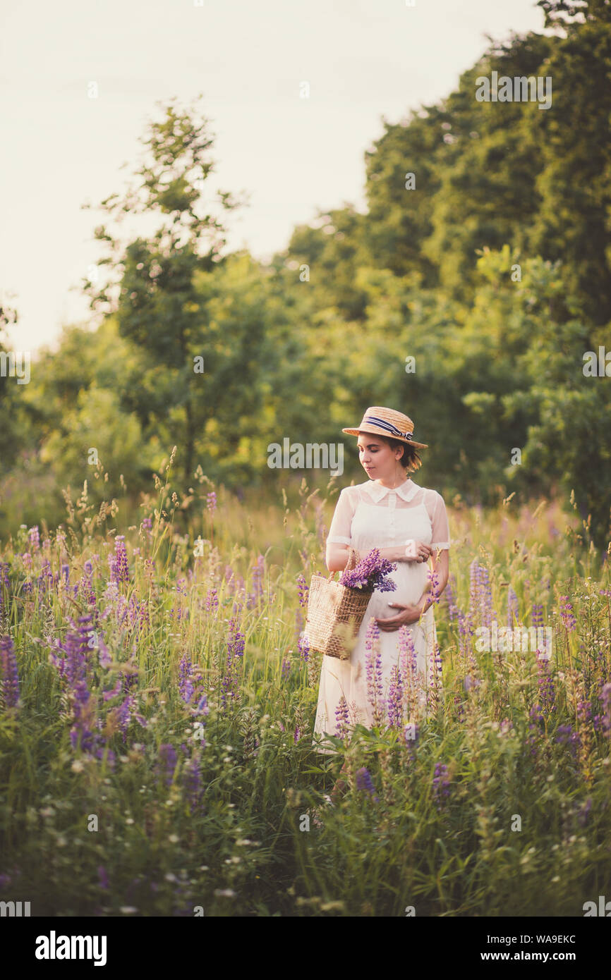 Donna incinta in abito bianco tenendo un cesto di paglia con fiori di campo Foto Stock