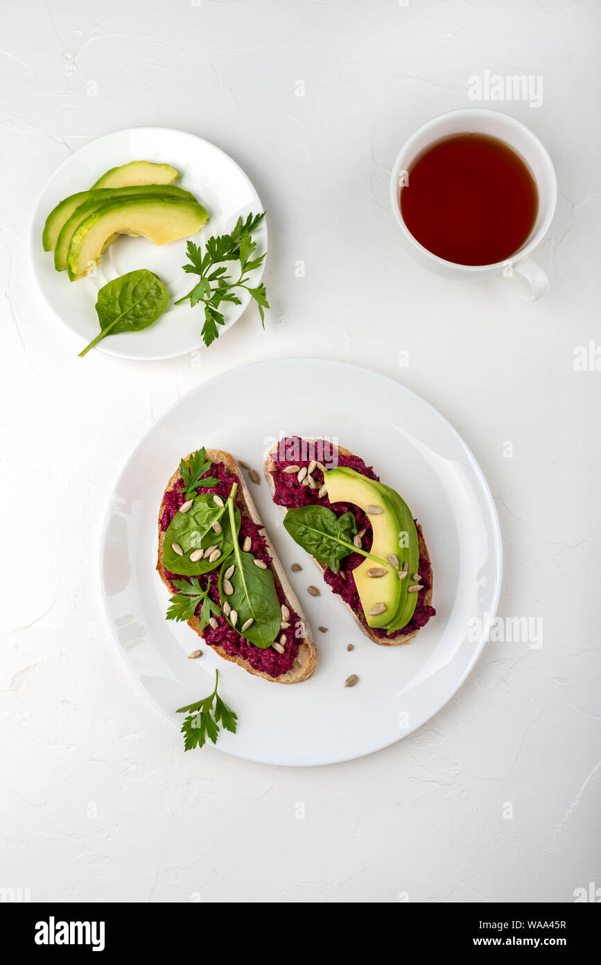 La prima colazione. Bruschette con barbabietola hummus, avocado, spinaci e semi su una piastra bianca e la tazza di tè. Vista dall'alto. Foto Stock