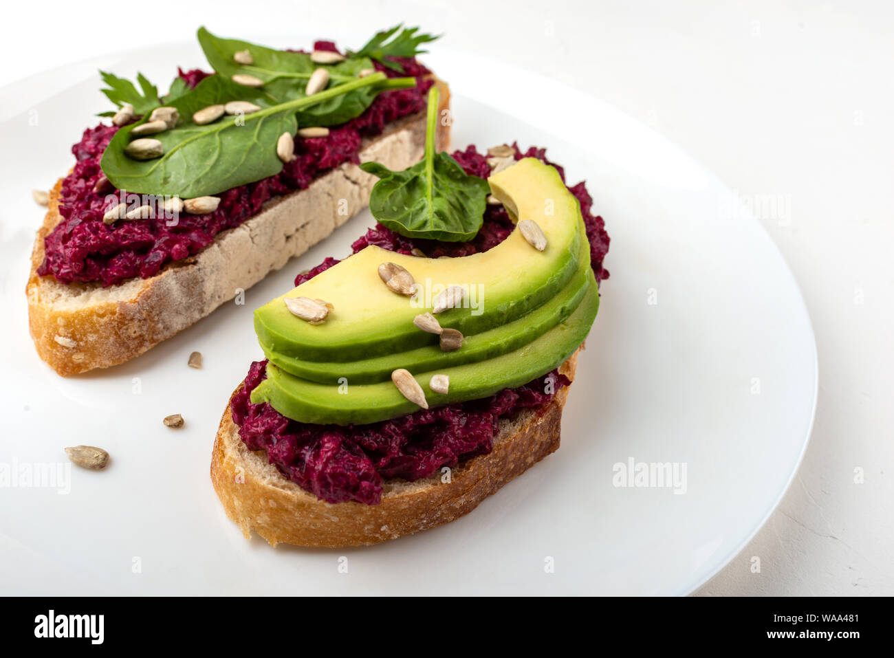 Bruschette con barbabietola hummus, avocado, spinaci e semi su una piastra bianca su sfondo bianco. Foto Stock