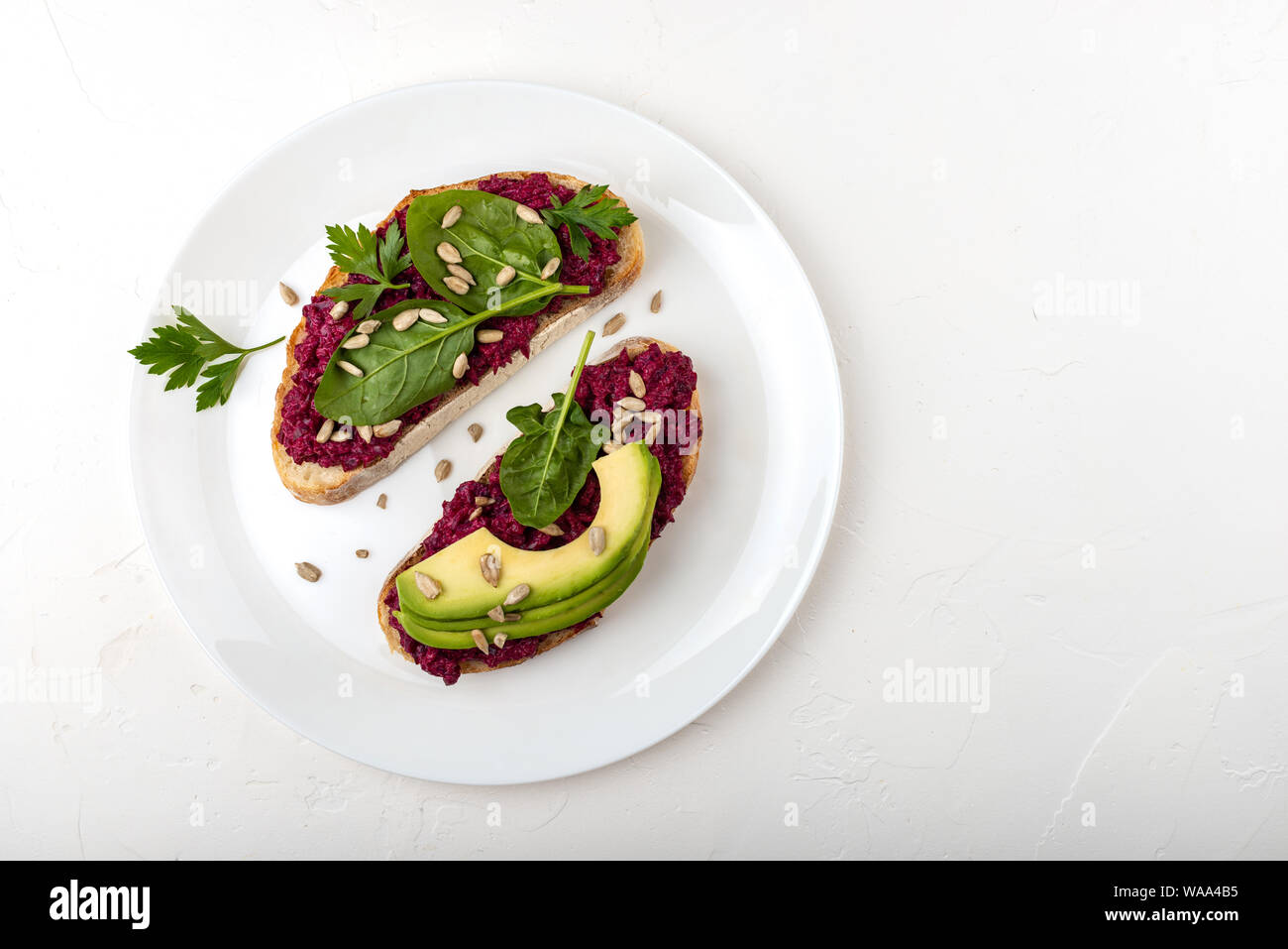 Bruschette con barbabietola hummus, avocado, spinaci e semi su una piastra bianca su sfondo bianco. Foto Stock
