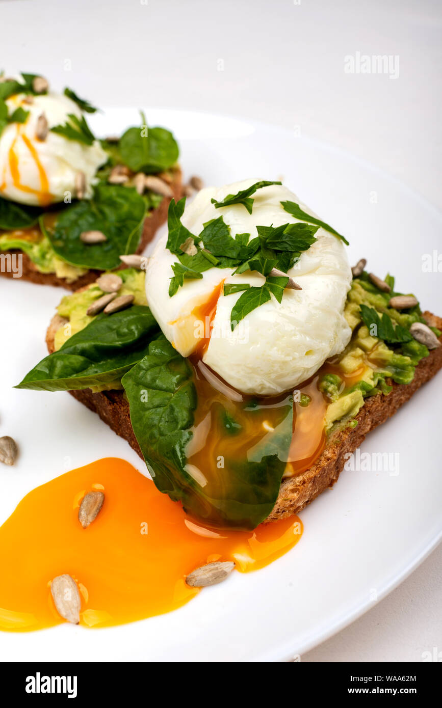 Uova al tegame con avocado, spinaci in foglie e semi su crostini di pane su una piastra bianca su sfondo bianco. Sana colazione o uno spuntino. Close-up. Foto Stock