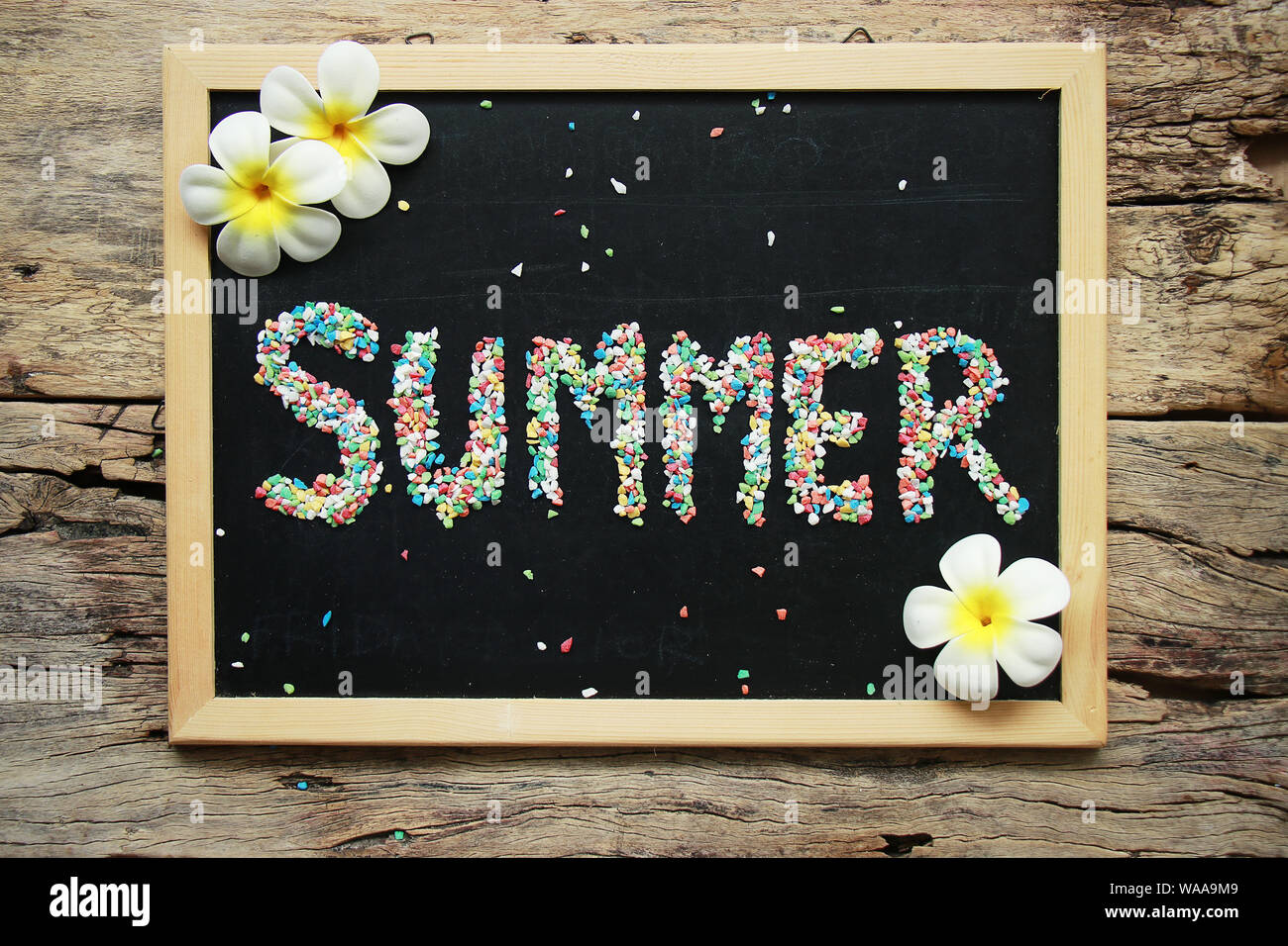Estate parola fatta da ghiaia colorata sul gesso nero bordo decorare con fiore tropicale su sfondo di legno Foto Stock