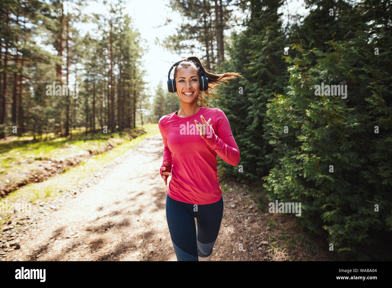 Giovani felici femminile ascoltare musica durante il jogging lungo un sentiero soleggiato nella foresta. Foto Stock