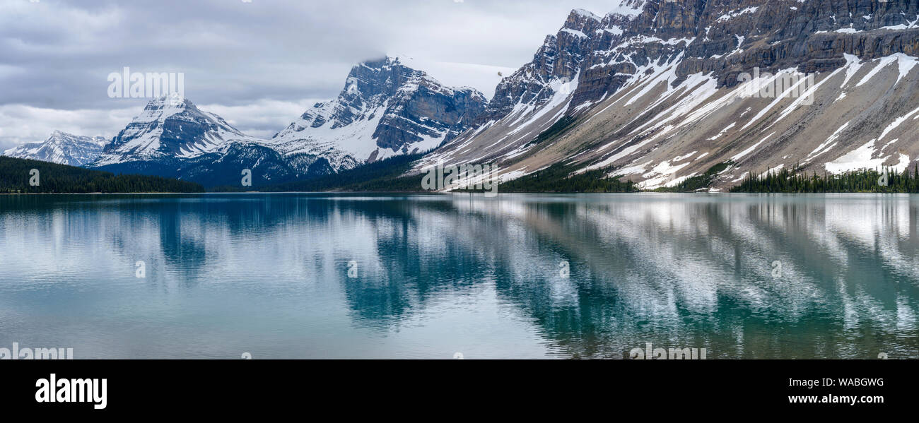 Al Lago Bow - Panoramico nuvoloso giorno di primavera vista delle cime innevate che riflette nella calma e colorata Bow Lake, il Parco Nazionale di Banff, AB, Canada. Foto Stock