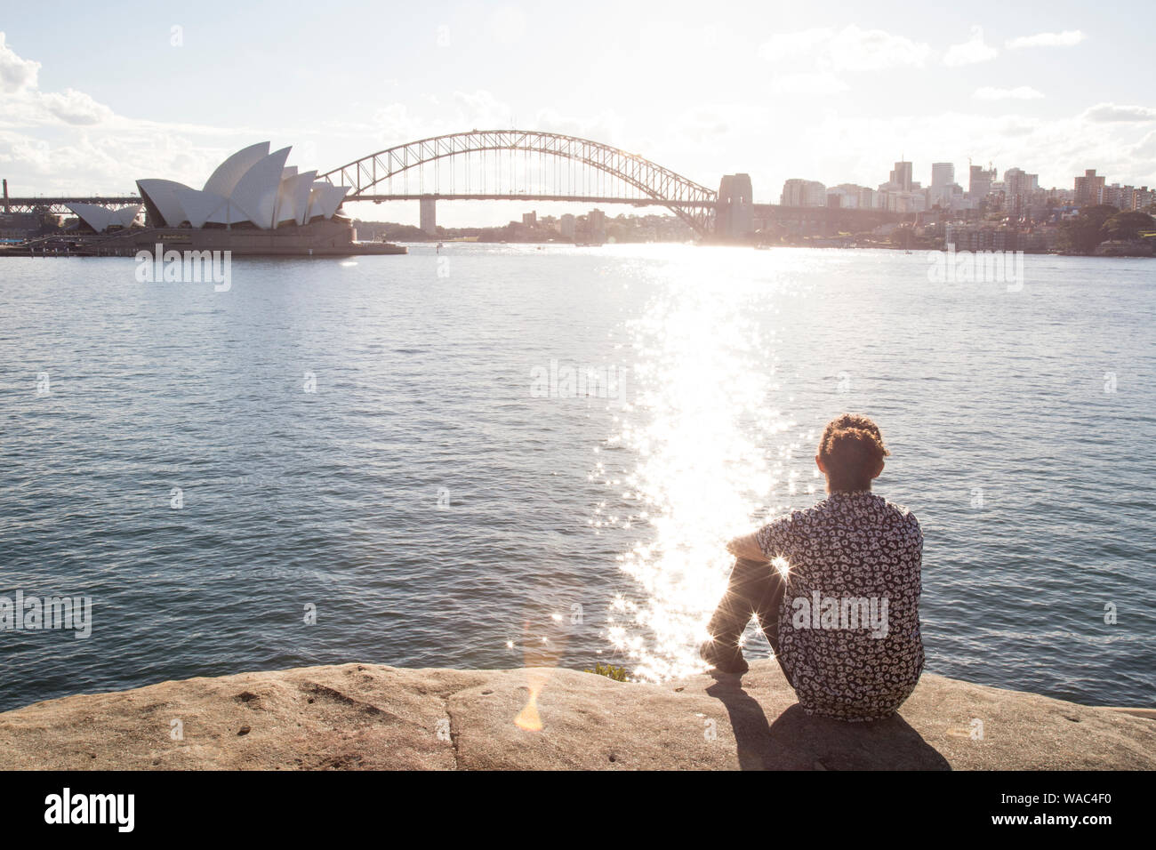 Uomo con bun, sitted sul bordo dell'acqua, fissando il Porto di Sydney Foto Stock