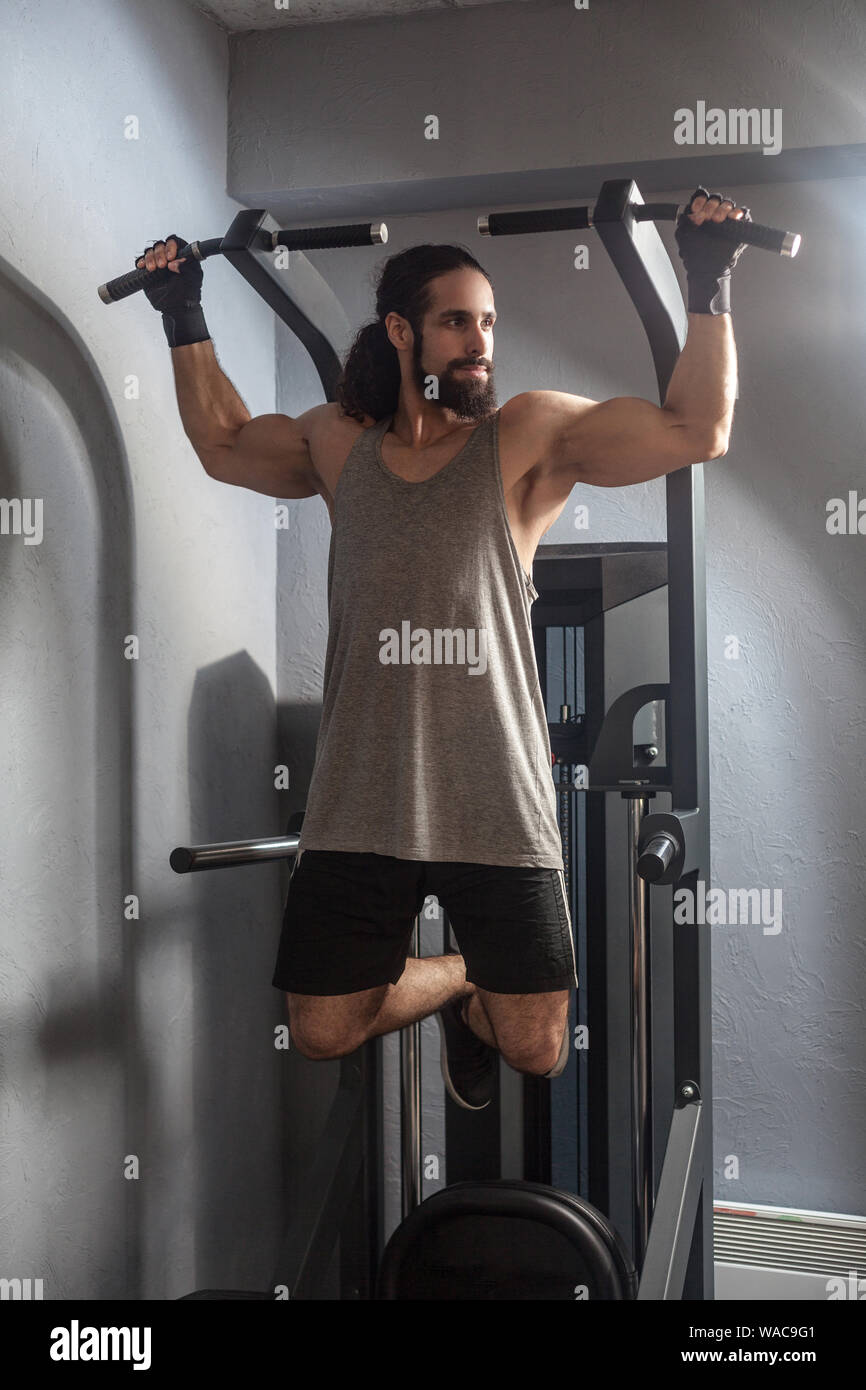 A piena lunghezza Ritratto di fiducioso giovane adulto uomo forte con lunghi capelli ricci che lavora fuori in palestra, appeso sulla barra orizzontale con le mani, facendo tirare ups Foto Stock